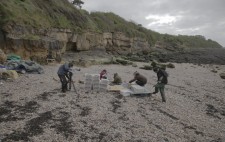 The beach is an expanse of shingle. Plastic crates are piled up next to a wooden pallet. A camera operator is filming as crew make ready the bonfire area.