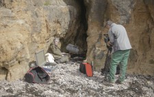 In the shelter of a hollow in the rocky cliff-face, Liz sits on beach stones, facing towards a large fluffy microphone on a stand. Nearby a camera operator sets up a camera on a tripod.