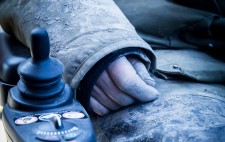 Liz hand rests on mud-coated trousers, next to the controls of a power wheelchair, her fingers caked in mud.