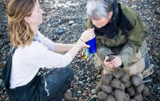 Liz drinks from through a staw, as her PA extends a blue water bottle. Liz holds a piece of clay, a heap of clay still remaining to be sculpted.