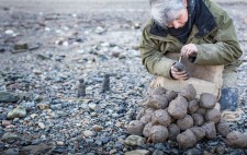 On a wide shingle foreshore, Liz leans over a pile of clay, sculpting a figure. To her side are two completed figures, which stands upon the shingle, almost camouflaged.