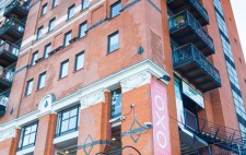 Liz is carried by three members of the team down a flight of metal steps. Towering above them is the red-brick Oxo building.