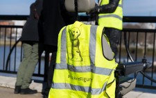 On the back of an empty wheelchair is a high vis jacket with the Figures logo, a drawn figure, and the text #We Are Figures. In the background, a member of the production team leans against the foreshore railing in conversation with audiences.