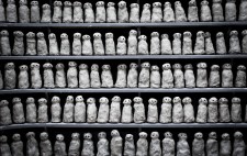 Over 100 small clay figures are lined up over five rows of shelving. Each is sculpted to the same design, yet varies in the detail, giving a sense of commonality and individuality.