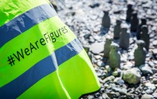 A high vis jacket with the project hashtag. In the background, 11 figures gather on the foreshore.