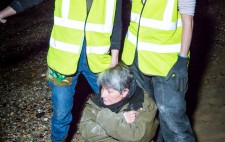 Liz sits in the sling on the foreshore, leaning against the legs of two members of the production team who stand behind her. All three look to a distant spot outside the picture frame.