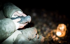 Liz holds a partially-sculpted figure in her hands, a mound of clay resting in front of her. In the background, a lit candle in a glass jar rests on the foreshore.