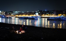 With a backdrop of city lights, two passersby sit on the foreshore facing Liz, in conversation, their faces illuminated by candlelight.