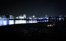 Against a deep black sky and a skyline of St Paul’s and the city, Liz sits with a few lanterns in front of her. She is a tiny figure in a dark landscape.