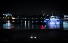 Seen from above, against a deep black sky and coloured lights reflected in the river, Liz sits with a few lanterns in front of her. She is a tiny figure, almost invisible against the dark of the foreshore.