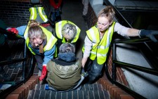 Liz is seated on a step in a sling, while three members of the production team get ready to lift her.