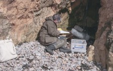 Facing into a hollow in the rocky cliff-face, Liz sits on beach stones, facing towards a large fluffy microphone on a stand and reading from an open folder.