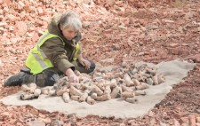 Liz sits in an expanse of crushed bricks, in reds and pinks, against a backdrop of woodland. She sifts through the fired figures, which are piled on a hessian mat, their colours echoing those of the landscape.