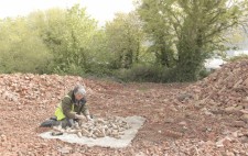 Liz sits in an expanse of crushed bricks, in reds and pinks, against a backdrop of woodland. She sifts through the fired figures, which are piled on a hessian mat, their colours echoing those of the landscape.