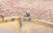 Liz sits in an expanse of crushed bricks, in reds and pinks. In the foreground is part of a yellow-painted industrial machine.