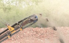 The arm of the machine projects upwards against a backdrop of brick and woodland. Small explosions of chips and dust fly into the air and downwards to ground.