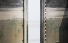 A gap opens between steel gates, the sea beyond. The image is horizontal strips of water and dry metal, sea and sky.