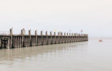 A wooden jetty, a stretch of vertical wooden planks, projects out to greys of water and sky, dwarfing a small sailing boat.