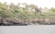 Looking back to shore from the boat, large houses are built at the top of a tree-lined cliff. A small group of people is gathered on the rocky cliff, looking out towards the boat.