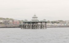 Rising from choppy water, with Clevedon seafront as a backdrop, is the wooden structure of the pier. The upper tier is surrounded by green railings with a pagoda at the centre.