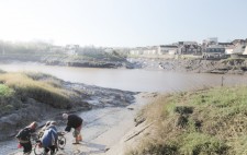 Liz is on a trike-wheelchair, tethered to a rope and assisted by three of the crew. The slipway opens out to wide banks of mud, the river beyond.