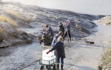 A wheeled cart with empty plastic crates is lowered down the slipway. Members of the team dig mud with spades from the riverbank.