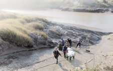 A wheeled cart with empty plastic crates sits at the bottom of the slipway. Members of the team dig mud with spades from the riverbank.