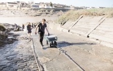 A wheeled cart with empty plastic crates is winched up the slipway. Below, members of the team dig mud with spades from the riverbank.