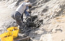 A member of the team digs with a space into a high bank of mud, two yellow tubs next to her, part-way to being filled.