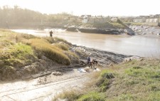 Members of the team dig the mud at the bottom of the slipway. On the grassy bank high above stands a camera operator at a tripod.