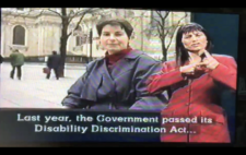 Screenshot from broadcast shows Liz Crow in medium shot to camera, with the Palace of Westminster in the background. Captions read 'Last year, the Government passed its Disability Discrimination Act...' To the right of screen is a BSL interpreter.