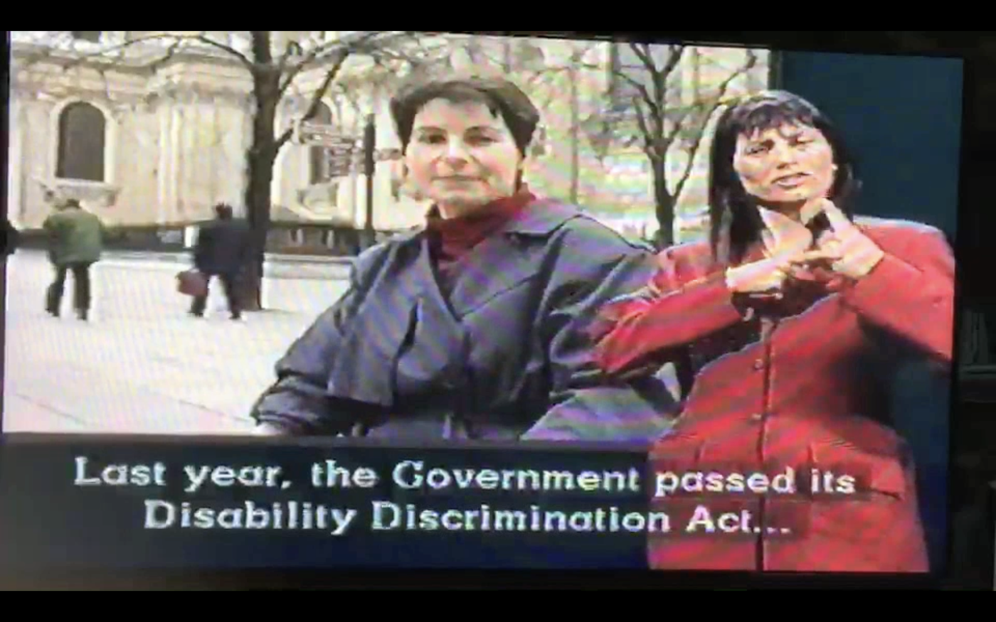 Screenshot from broadcast shows Liz Crow in medium shot to camera, with the Palace of Westminster in the background. Captions read 'Last year, the Government passed its Disability Discrimination Act...' To the right of screen is a BSL interpreter.