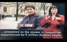 Screenshot from broadcast shows Liz Crow in medium shot to camera, with the Palace of Westminster in the background. Captions read '...presented as the answer to inequalities experienced by 6 million disabled people' To the right of screen is a BSL interpreter.