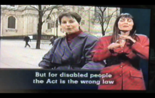 Screenshot from broadcast shows Liz Crow in medium shot to camera, with the Palace of Westminster in the background. Captions read 'But for disabled people the Act is the wrong law'. To the right of screen is a BSL interpreter.